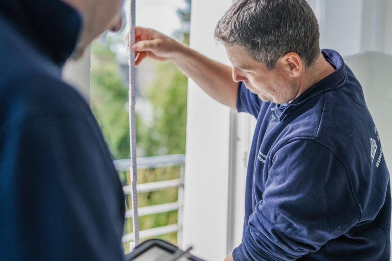 Zwei Mitarbeiter von Fensterbau Knipping nehmen präzise Fenstermaße – einer misst mit einem Zollstock, der andere notiert die Daten digital auf einem Tablet.