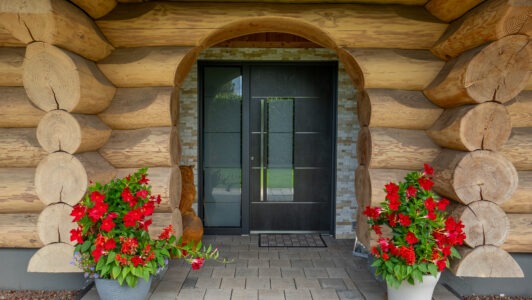 Moderne, dunkle Haustür mit Glaseinsatz in einem Blockhaus mit markanten Rundholzstämmen und dekorativen Blumenkübeln am Eingangsbereich.