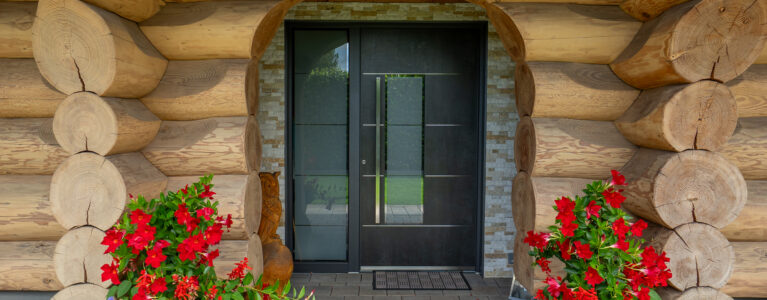 Moderne, dunkle Haustür mit Glaseinsatz in einem Blockhaus mit markanten Rundholzstämmen und dekorativen Blumenkübeln am Eingangsbereich.