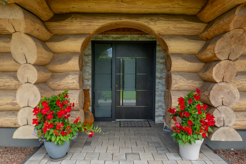 Moderne, dunkle Haustür mit Glaseinsatz in einem Blockhaus mit markanten Rundholzstämmen und dekorativen Blumenkübeln am Eingangsbereich.