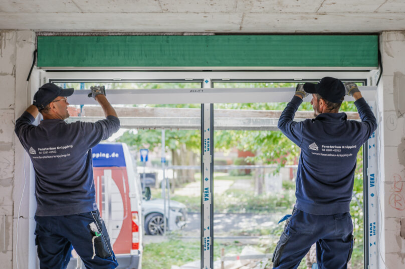 Zwei Monteure von Fensterbau Knipping setzen gemeinsam ein neues Fenster in einen Rohbau ein, mit Blick nach außen auf den Firmenwagen.