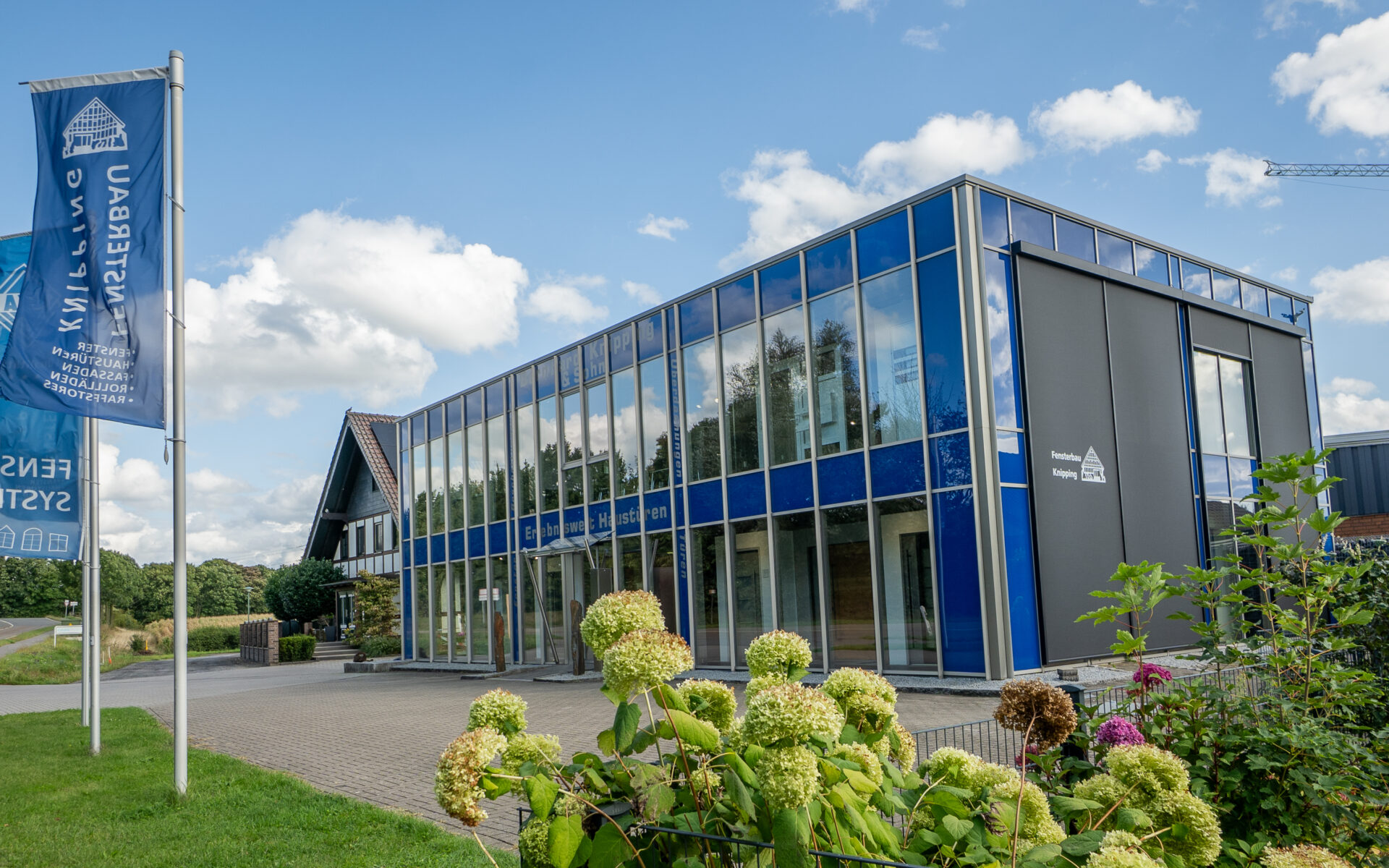 Modernes Firmengebäude von Fensterbau Knipping mit Glasfassade, blauen Firmenfahnen und gepflegtem Außenbereich bei sonnigem Wetter.