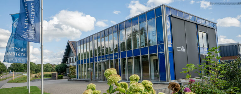 Modernes Firmengebäude von Fensterbau Knipping mit Glasfassade, blauen Firmenfahnen und gepflegtem Außenbereich bei sonnigem Wetter.