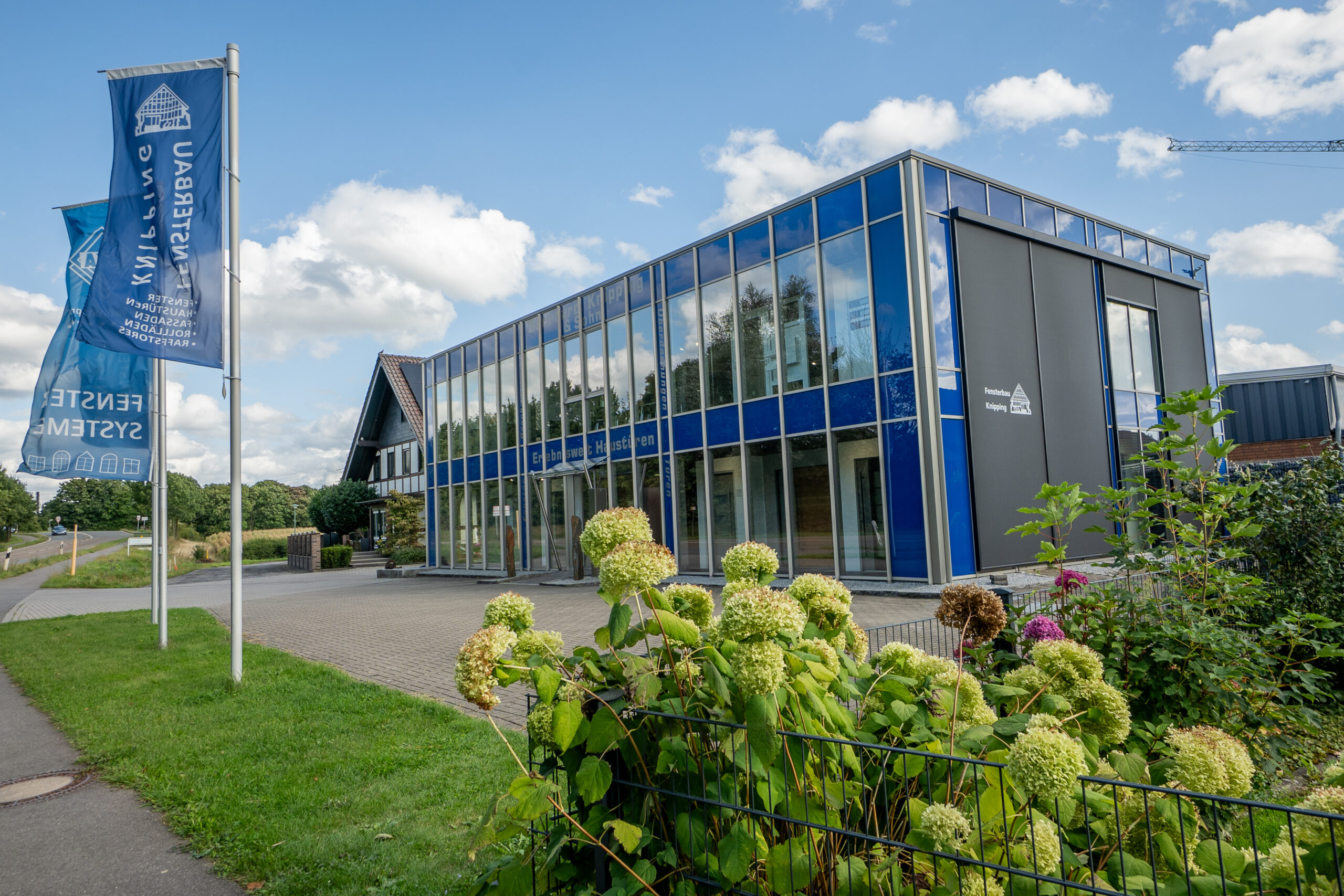Modernes Firmengebäude von Fensterbau Knipping mit Glasfassade, blauen Firmenfahnen und gepflegtem Außenbereich bei sonnigem Wetter.